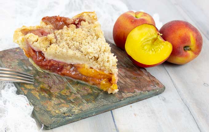 slice of Fresh Peach Pie With Crumb Topping on slate with whole peaches