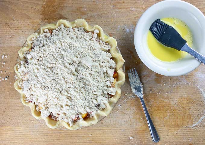 uncooked Fresh Peach Pie With Crumb Topping with bowl of egg wash
