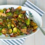 Roasted Brussels Sprouts with Bacon and Shallots on white oval dish on whitewashed plank background with napkin and spoon