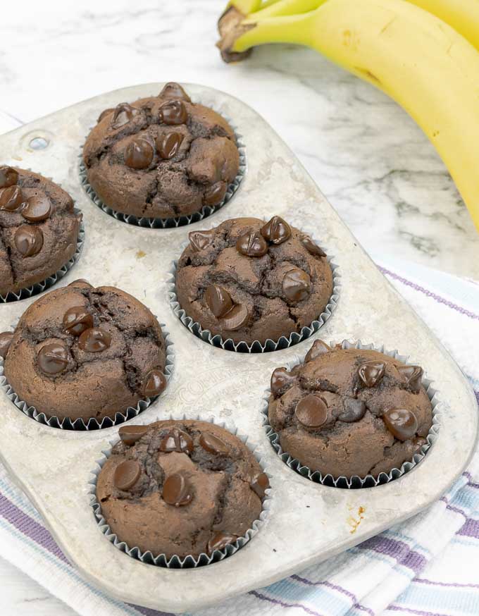 six double chocolate banana muffins in a metal muffin tin next to bananas