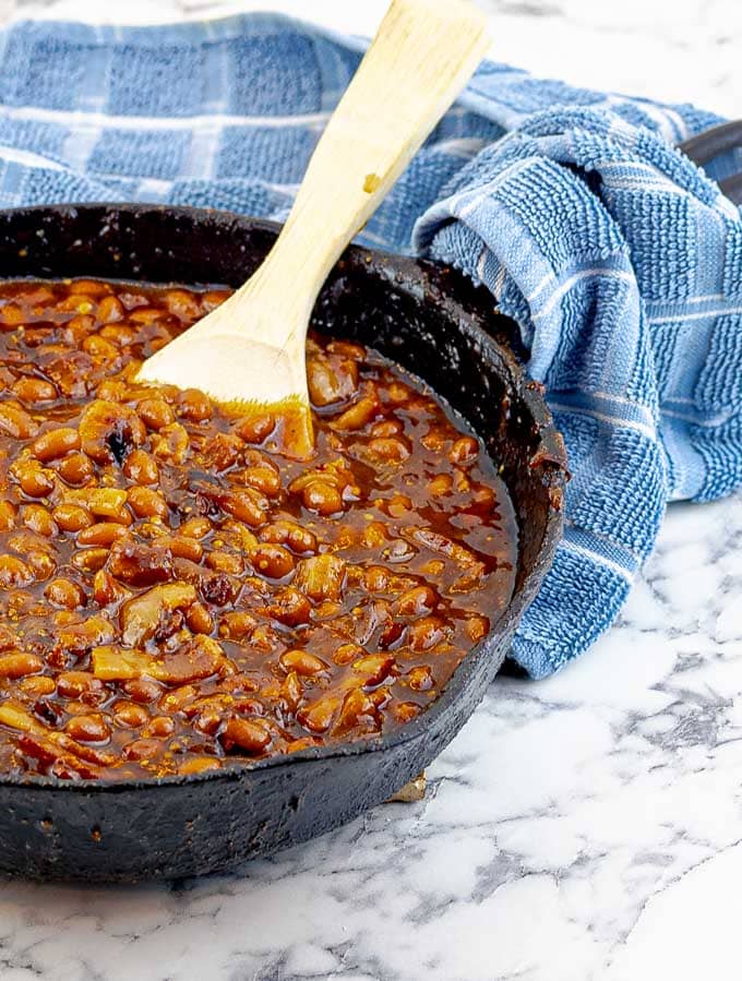 Bourbon Bacon Grilled Baked Beans in a cast iron skillet with a wooden spoon and blue towel