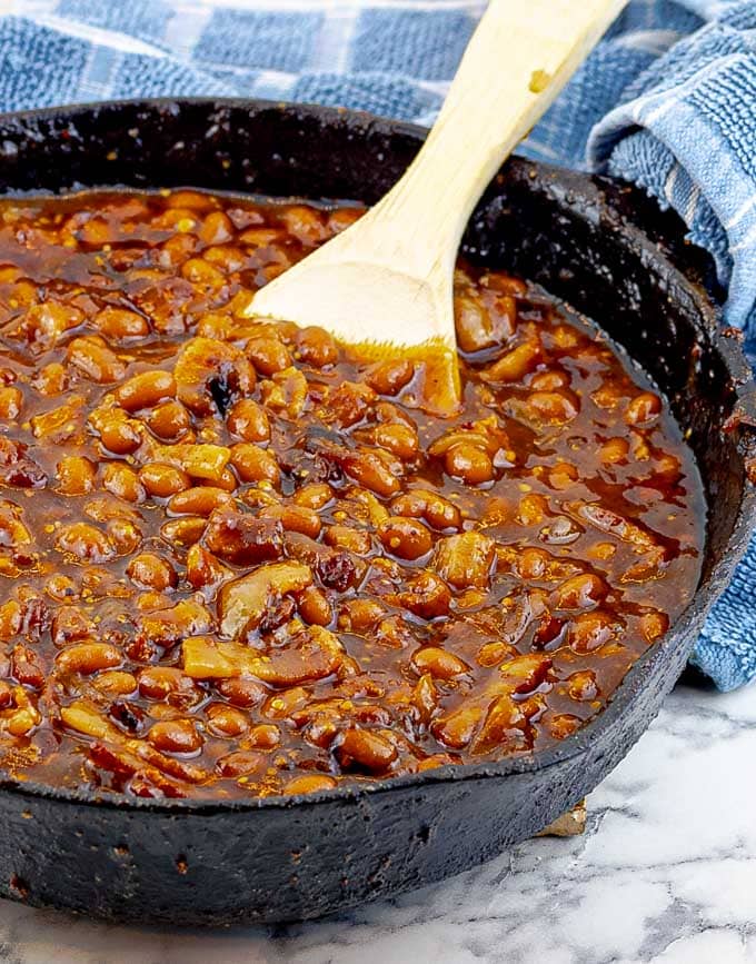 Bourbon Bacon Grilled Baked Beans in a cast iron skillet with a wooden spoon and blue towel