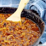 Bourbon Bacon Grilled Baked Beans in a cast iron skillet with a wooden spoon and blue towel
