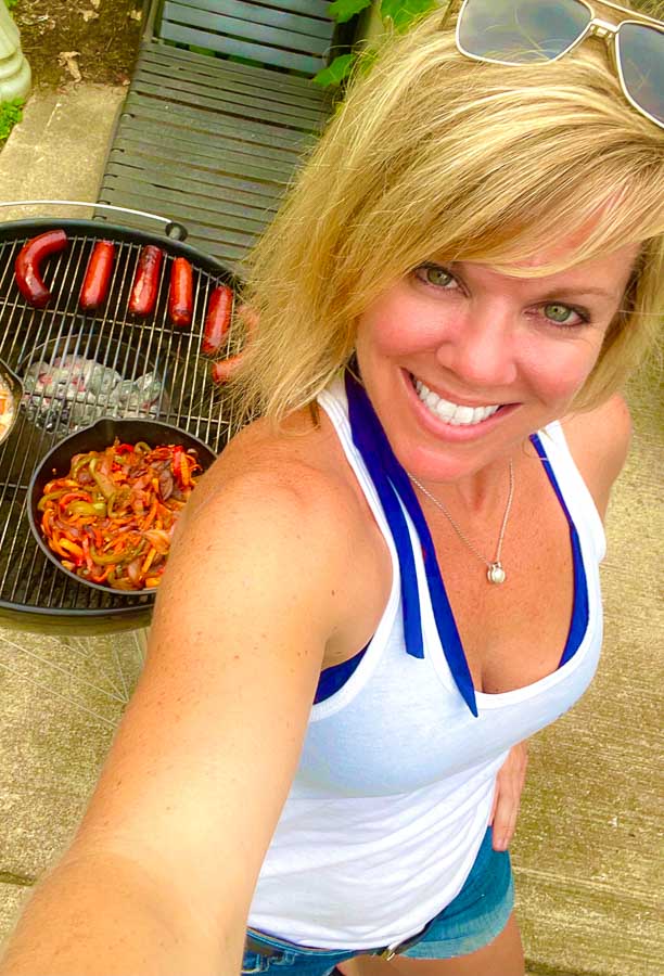 Jennifer with weber grill behind grilling sausages, onions, and peppers