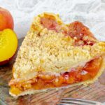 piece of Fresh Peach Pie With Crumb Topping next to a sliced peach