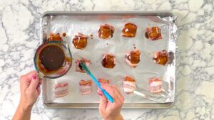 sausages wrapped in bacon getting a brown sugar glaze on a foil lined baking sheet